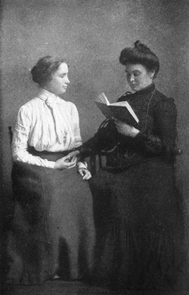 Anne Sullivan is reading out of a book an spelling the words into Helen&#x27;s hand